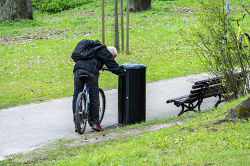 Elderly Man Searching Trash Can in City Park While Sitting on Bicycle, Concept of Poverty and...