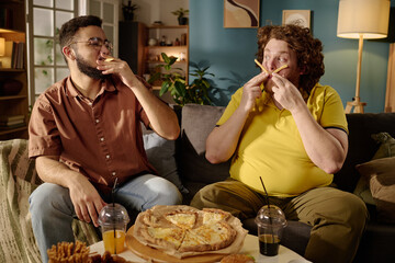 Two men sitting on couch eating pizza and snacks while having fun. Room is warmly lit with comfortable ambiance and various decorations creating cozy atmosphere