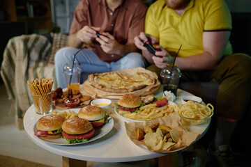 Two friends sitting and enjoying a fun gaming session with a large spread of delicious fast food on...