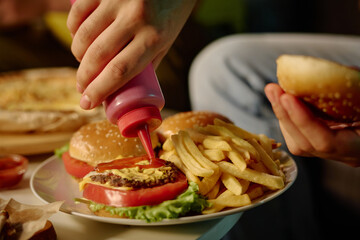 A close-up of persons hands adding sauces on homemade burger with fresh lettuce and fries in background creating a mouth-watering scene