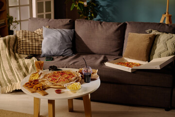 Inviting living room setting featuring a sofa with cushions and throw blankets. Coffee table with pizza, snacks, and drinks set for cozy evening in relaxed atmosphere