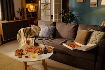 Sofa with pillows and blankets in a cozy living room, table with pizza, snacks, and drinks, creating a comfortable atmosphere perfect for relaxing or socializing