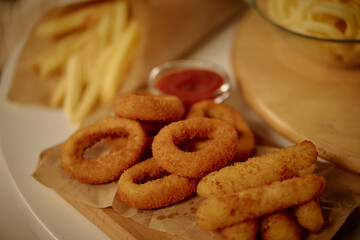 Plate of crispy onion rings, French fries, and mozzarella sticks arranged on parchment paper with a side of ketchup on a wooden board in cozy setting