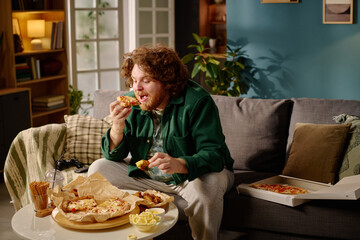 Young man sitting on sofa, enjoying a slice of pizza with cozy and relaxed surroundings. Various...