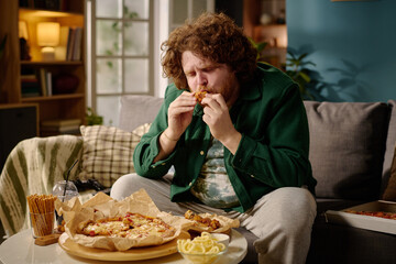 Man sitting on couch enjoying pizza with other snacks on table in front of him, surrounding him with cozy living room decor