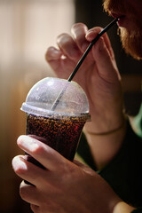 Close-up of person's hand holding cold drink in plastic cup with straw. Side profile of bearded...