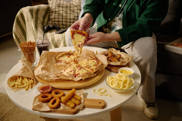 Person grabbing slice of pizza from table filled with various snacks and drinks while relaxing on sofa in cozy living room. Cheerful dining experience with comfort food and beverages