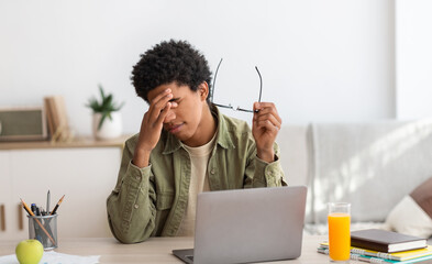 Black teenager feeling tired, rubbing irritated eyes, sitting at desk with laptop, exhausted from...