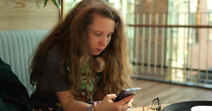 Teenage girl with headphones around neck types on smartphone waiting for order on cafe terrace. Child fingers clack away on phone navigating social media