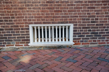 square wooden vent in the wall of an old brick house by the brick sidewalk.