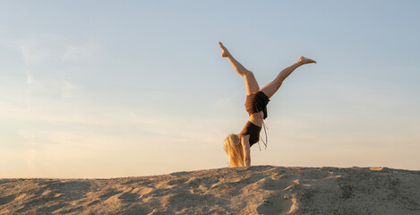 A woman showcases her balance with a handstand on a sandy dune in the desert during sunset,...
