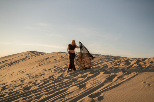 A woman stands in the desert, holding a light fabric poreo as the sun sets, casting long shadows over the sand dunes around her.