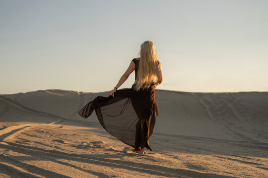 A woman stands in the desert, holding a light fabric poreo as the sun sets, casting long shadows over the sand dunes around her.