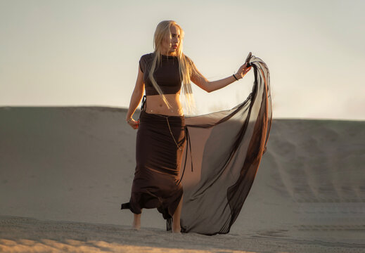 A woman stands in the desert, holding a light fabric poreo as the sun sets, casting long shadows over the sand dunes around her.