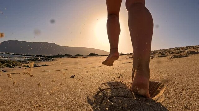 LOW ANGLE VIEW, SLOW MOTION, LENS FLARE: Sporty woman on an evening run along a sandy ocean shore. At sunset, she runs barefoot on beach in blue sportswear, leaving flying sand behind in golden light.