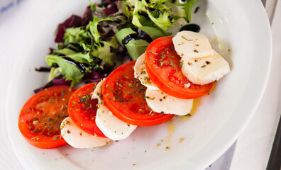 Mediterranean food, caprese salad - tomatoes, mozzarella cheese, dried basil and greens with balsamic on white plate