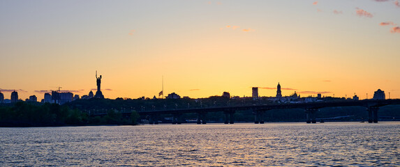 Fototapeta premium Beautiful sunset over Kyiv city skyline with Dnipro River, Paton Bridge, and Motherland Monument silhouette under colorful sky
