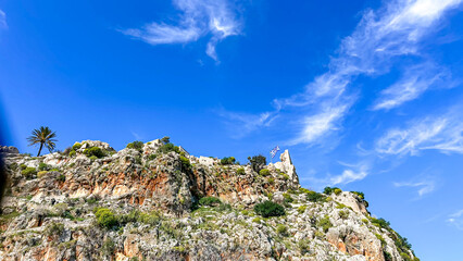 Naklejka premium Ancient Stone Ruins With Greek Flag On Rocky Hilltop In Greece Under Bright Blue Sky, Concept Of Heritage, Summer Travel, And Historical Tourism