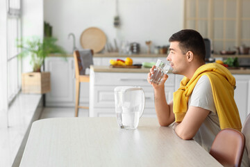 Young man with filter jug drinking fresh water at table in kitchen