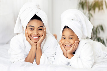 Beauty Portrait Of Happy Black Mom And Daughter In Bathrobes And Towels On Head Lying On Bed At Home, Posing After Bath, Having Spa Day, Cute Little Girl And Her Mother Having Fun Together, Free Space