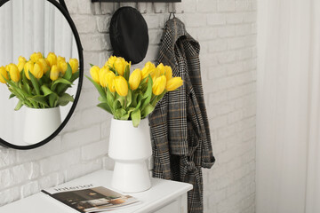 Vase of yellow tulips on dresser with mirror, hanger and coat near white brick wall in hallway
