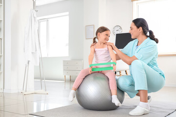 Female osteopath and little patient with elastic band doing exercises on fit ball in clinic