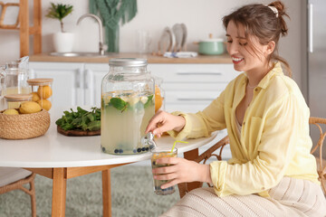 Young woman pouring lemonade from jar into glass on table in kitchen
