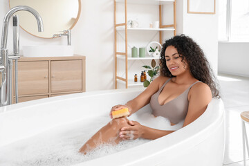 Beautiful young happy African-American woman taking bath with sponge and soap foam at home