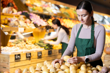Adult woman seller in apron puts potatoes on display in vegetable shop