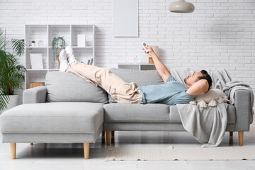Young man with air conditioner remote control lying on sofa near white brick wall