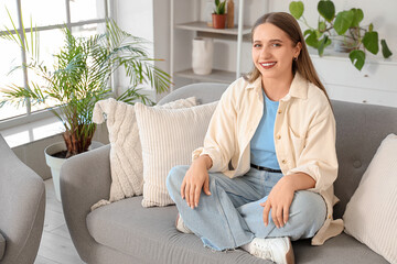 Fototapeta premium Smiling young woman relaxing comfortably on a gray sofa in a bright modern living room with houseplants