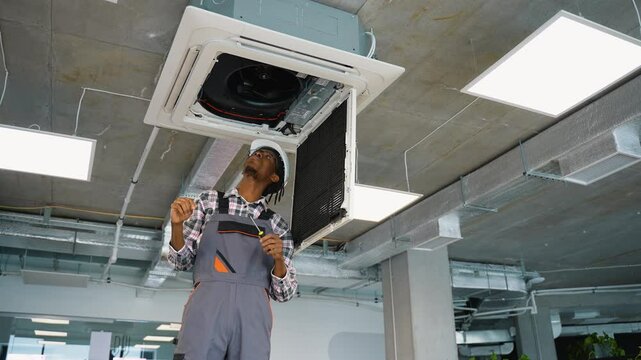 African American maintenance worker in uniform and helmet repairs a ceiling air conditioning unit