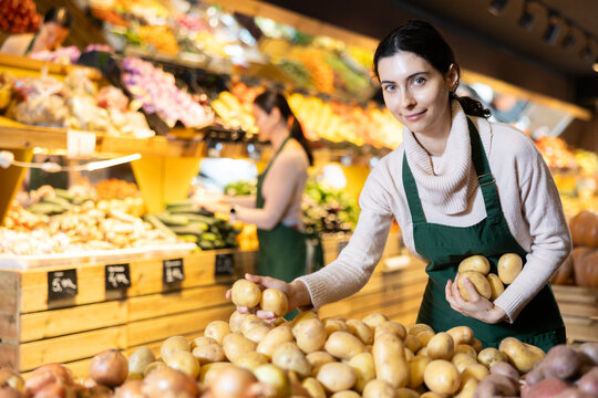 Female employee informs about arrival of ripe potato, advertises product. Assistant seller in apron, forms display case in vegetable store. Local and imported farm vegetables and fruits.