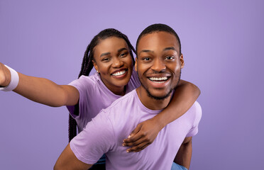 Obraz premium Having Fun. Portrait of happy black couple talking selfie, cheerful woman piggyback riding on her boyfriend's back, holding looking at camera standing isolated over purple studio wall