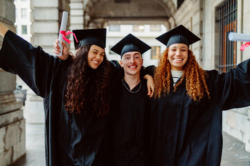 Three cheerful students wearing graduation gowns and mortarboards are holding their diplomas and...