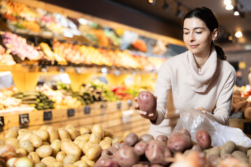 Near shelves and display cases in sales area of store, woman customer takes ripe potato out of box. Wide range of agricultural products. Self-service, comfortable shopping.