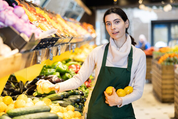 Vegetable shop assistant working in greengrocery. Woman seller in sales area with lemon in hands. Employee demonstrates supply of fruit. Local and imported farm vegetables and fruits
