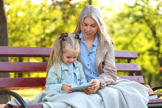 Cute little girl and her mother with warm plaid using tablet computer on bench in park