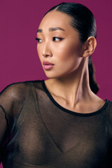 Asian woman in sheer top posing against purple background with hands on hips and hair in ponytail