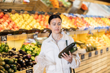Portrait of female shopper carefully selecting ripe zucchini in supermarket