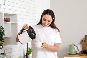 Beautiful young happy woman pouring hot water from modern electric kettle into cup in kitchen