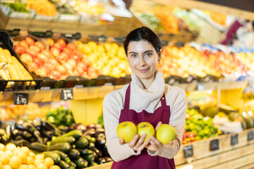 Young woman seller in apron offers fresh apples in vegetable shop