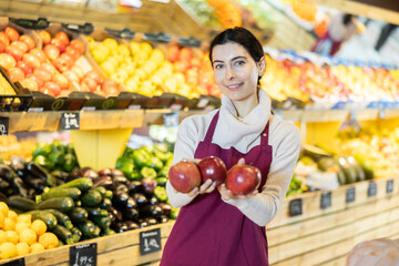 Female saleswoman offers ripe red apples in a supermarket. Employee of a vegetable store invites you to buy seasonal fruits