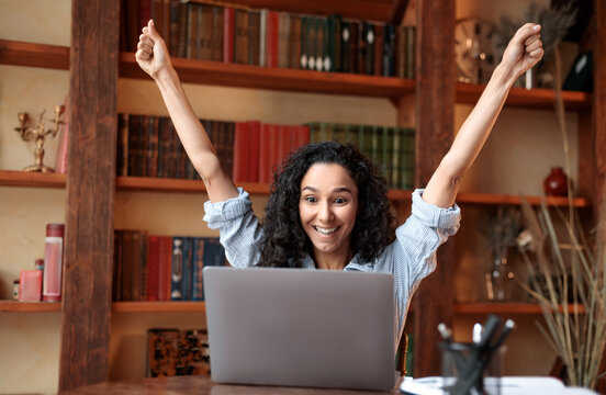Yes, I'm A Winner. Excited emotional ecstatic lady celebrating success, victory or great news sitting at table, using personal computer, screaming yeah and raising hands up. Smiling happy woman in joy