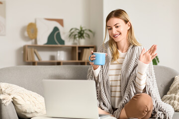 Fototapeta premium Young woman with cup of tea and laptop video chatting on sofa at home