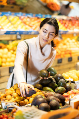 Young woman buyer choosing fresh avocados in vegetable shop