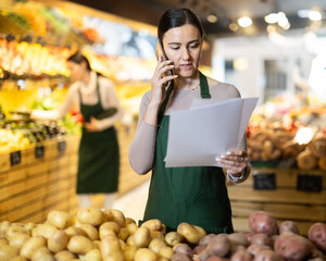 Adult woman seller takes inventory with checklists and talks on mobile phone in vegetable shop