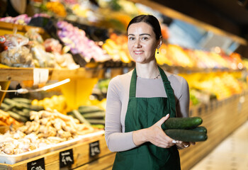 Polite middle-aged female seller offering cucumber standing by counter in large grocery store