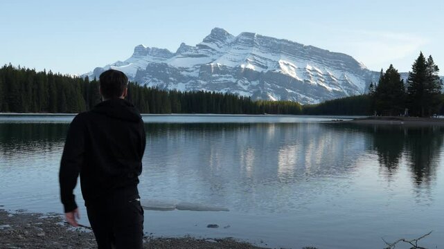 Young Male hiker throwing rocks on a lake. Happy man enjoying Mount Rundle scenery and nature and calm , Banff National Park. adult male skimming pebbles on a water of Two Jack Lake surface - 4K Video