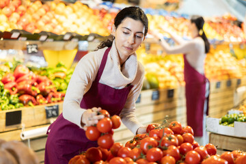 Female seller stands near the counter with red and tomatoes, checks the quality and puts them on the shelf. Supermarket worker keeps an eye on the windows